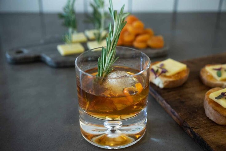 A smoked Christmas Manhattan drink with a large ice cube, orange peel and rosemary sprig