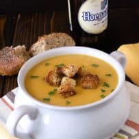 A white soup bowl with handles filled with beer soup and croutons with crusty bread and a bottle of beer in the background