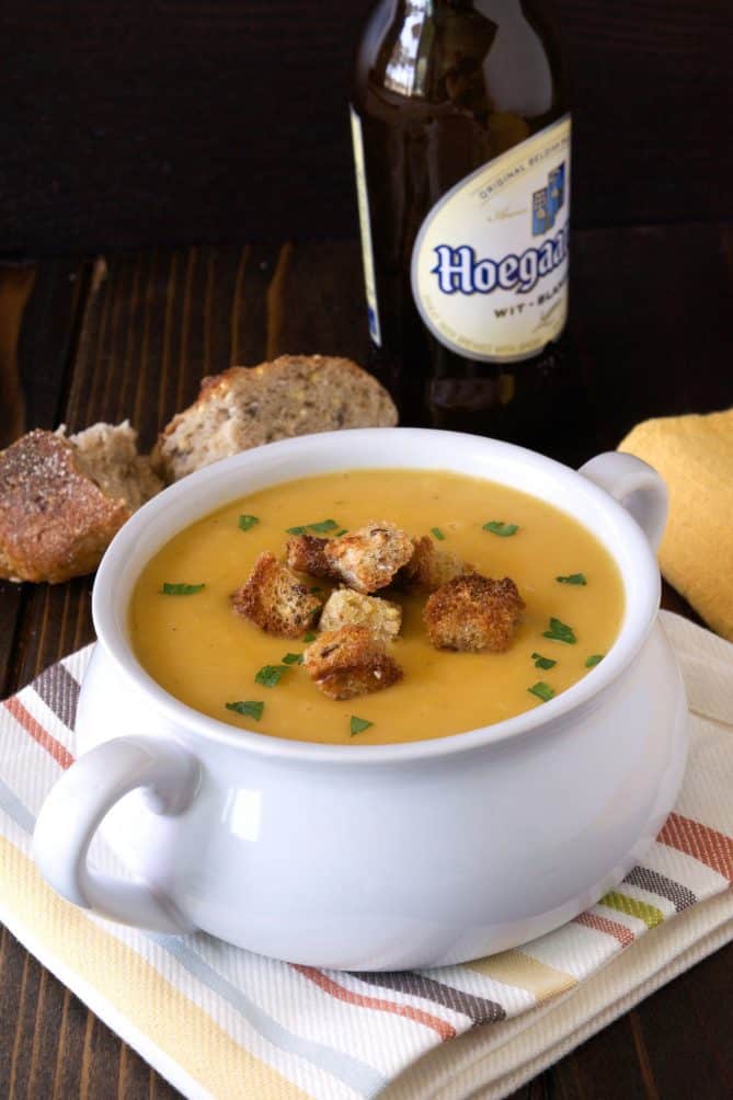 A white soup bowl with handles filled with beer soup and croutons with crusty bread and a bottle of beer in the background