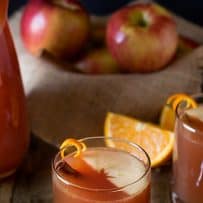 Dark orange colored apple punch in a glass with half an apple slice floating on top with orange twist and cinnamon stick