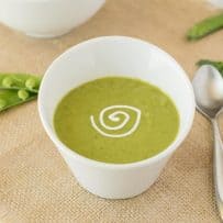 Vibrant green pea soup in a white garnished with sour cream with a spoon and fresh pea pods