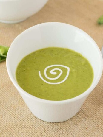 Vibrant green pea soup in a white garnished with sour cream with a spoon and fresh pea pods