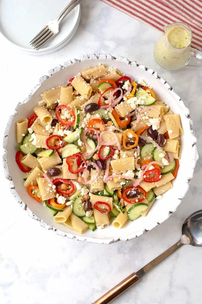 Summer Greek pasta salad from overhead in a white bowl with dressing, a serving spoon and a fork