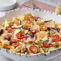 A large white bowl filled with pasta, colorful vegetables, oilves and cheese