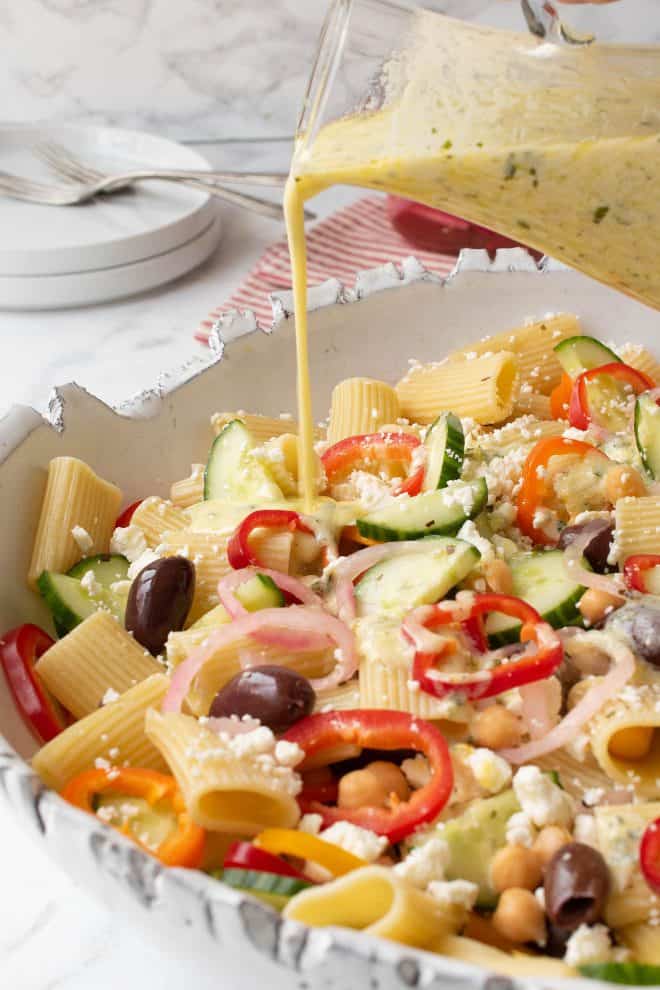 The yummiest and creamy dressing being drizzled over a summer Greek pasta salad