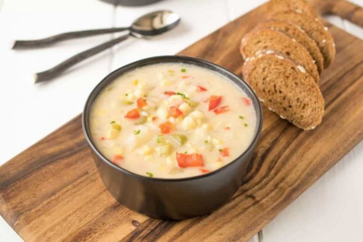 A black bowl filled with corn chowder and red peppers on a board with sliced bread