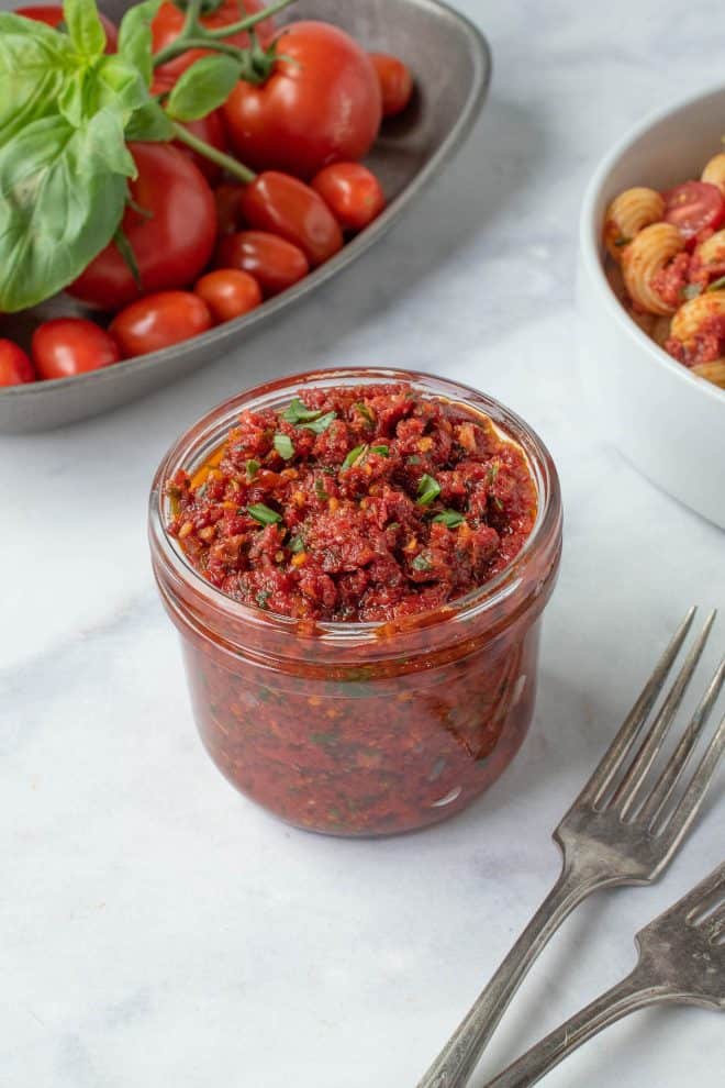 A jar of sun-dried tomato pesto with a bowl of pasta and fresh tomatoes and basil