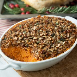 A white oval casserole dish filled with sweet potato casserole with pecan topping with a turkey in the background