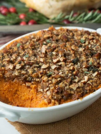 A white oval casserole dish filled with sweet potato casserole with pecan topping with a turkey in the background