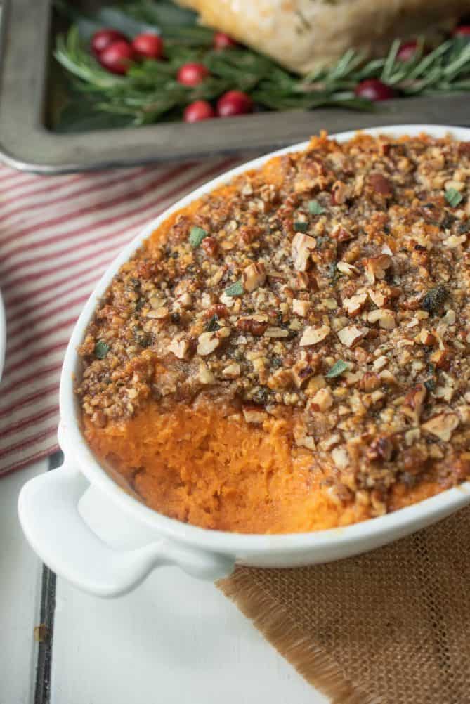 A closeup of a sweet potato casserole with a scoop taken out showing the chopped pecan topping flecked with fresh sage