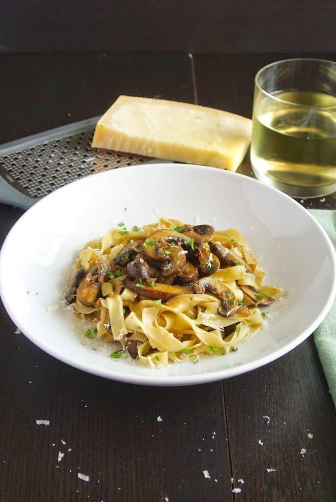 A large white bowl of tagliatelle with mushrooms with a glass of wine and Parmesan cheese