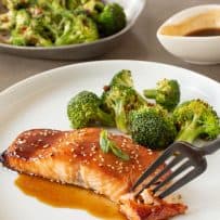 Using a fork to get a bite of teriyaki glazed salmon on a white plate with broccoli