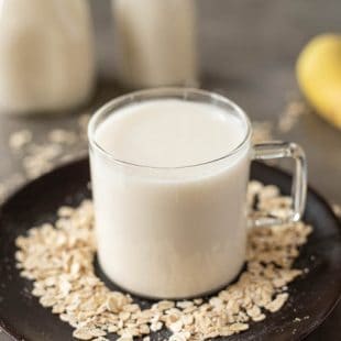 Oat milk in a glass mug on a dark plate with bottles of oat milk and a banana