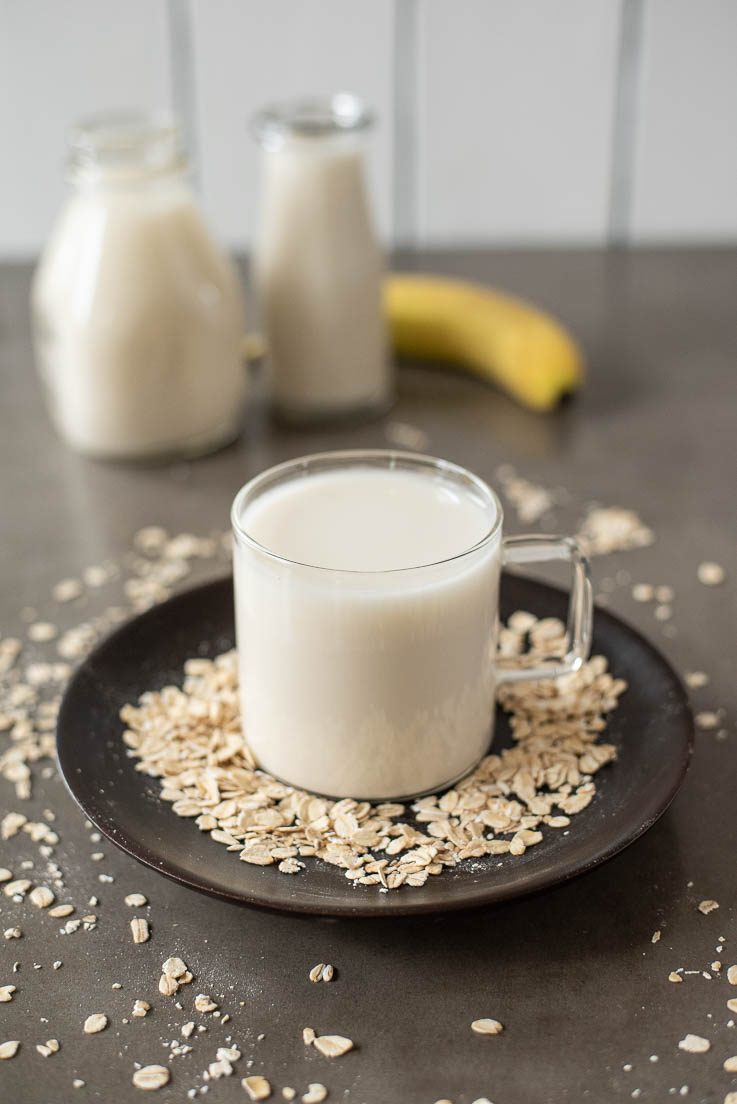 A glass mug filled with oat milk on a dark plate surrounded by old fashioned oats