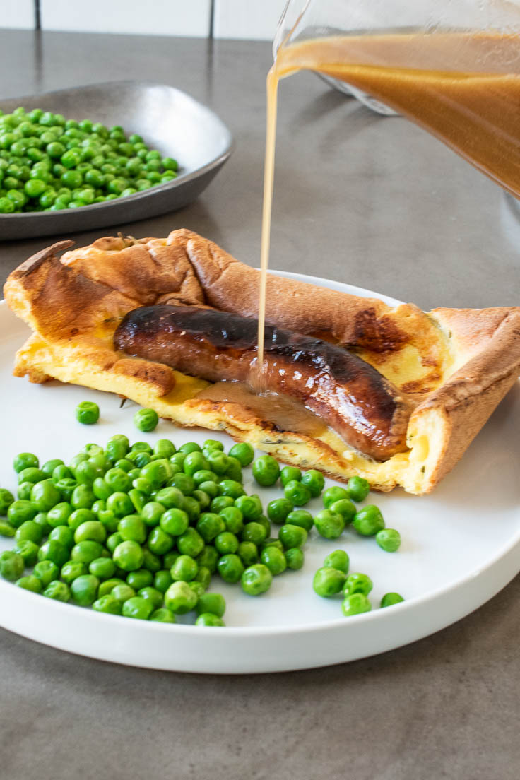 Gravy being poured over a piece of toad in the hole on a white plate with peas