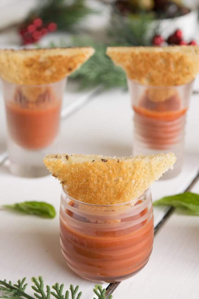 A closeup of a shot glass of tomato soup with a mini grilled cheese triangle on top