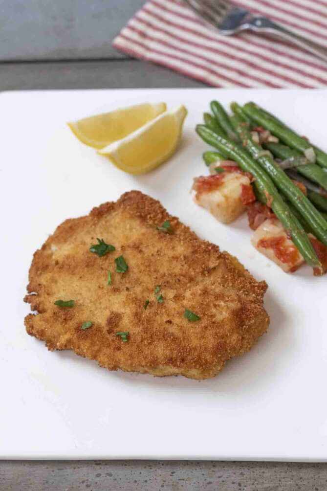 A chicken cutlet on a square plate with green beans and lemon wedges.