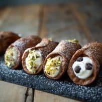 Cannoli lined up on a slate board, with chopped pistachios and chocolate chips