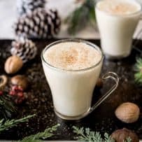 Eggnog in glass mugs surrounded by shelled nuts, pine cones and fern