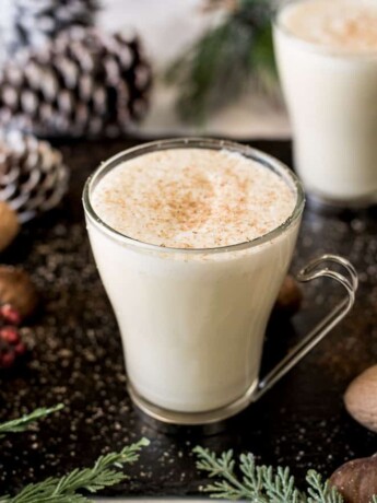 Eggnog in glass mugs surrounded by shelled nuts, pine cones and fern