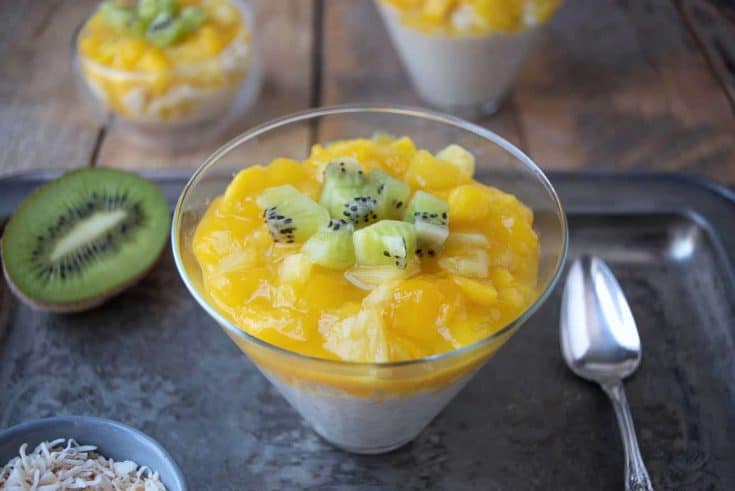 Tropical rice pudding in a coned shape glass topped with fresh mango, kiwi and pineapple with a spoon and extra kiwi