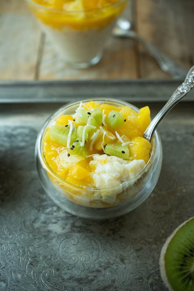A glass bowl with a spoon in rice pudding with mango, pineapple and kiwi