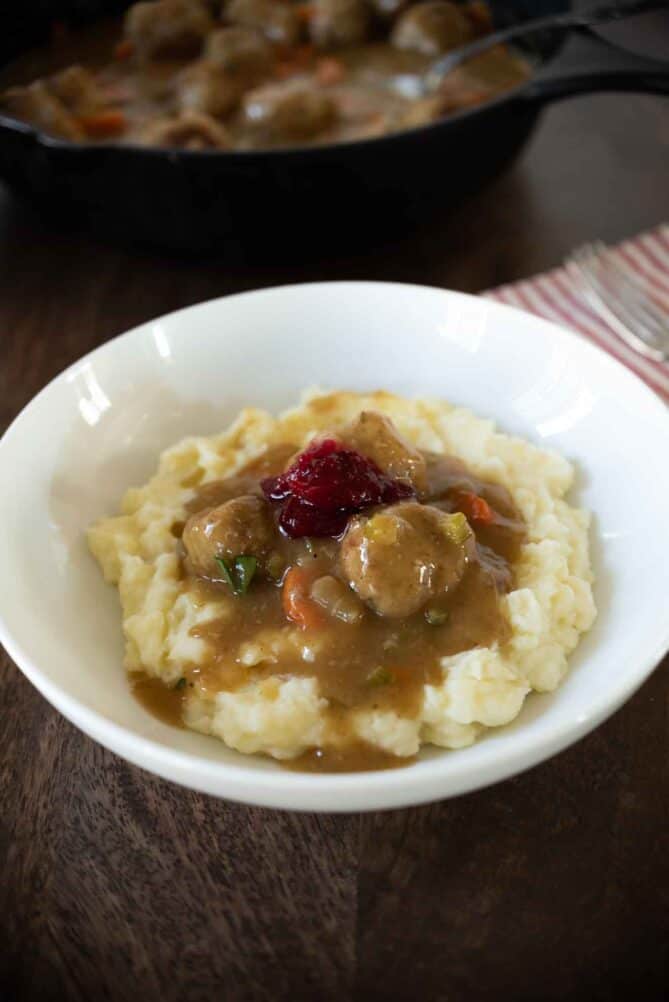A white plate of mashed potatoes topped with turkey meatballs, gravy and a little cranberry sauce