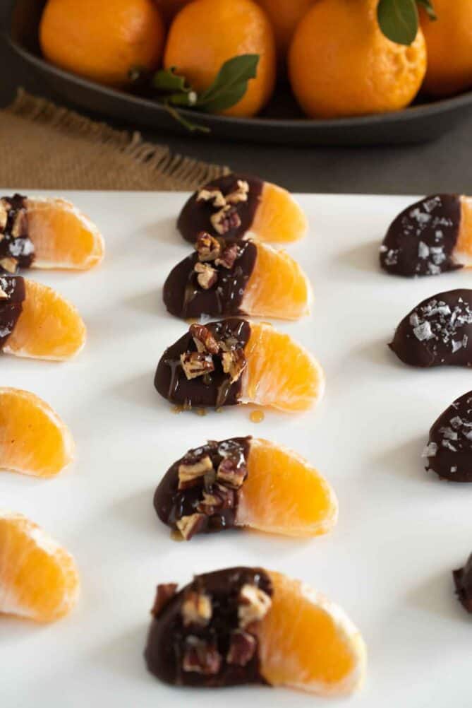 Orange segments lined up on a white plate that are dipped in melted chocolate with pecans, caramel and sea salt