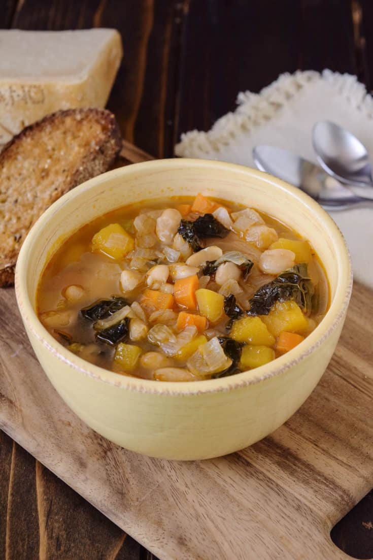An off white bowl filled with Tuscan bean soup with bread, Parmesan and spoons