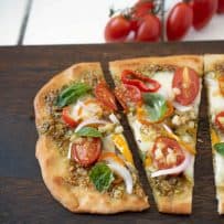 A closeup of a slice of vegetable pesto flatbread pizza showing the cherry tomatoes, red onion, corn, peppers, pesto and fresh basil