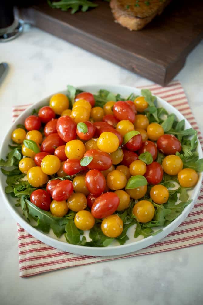 A white serving dish with fresh arugula topped with warm cherry tomato and basil salad using red and yellow tomatoes