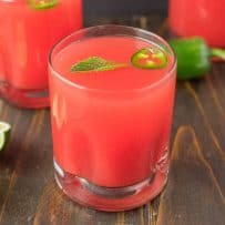 A vibrant watermelon colored drink with green mint and jalapeño slice floating on top