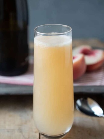 Prosecco and peach puree mixed in a stemless tall Champagne glass with a bottle of Prosecco and peach halves