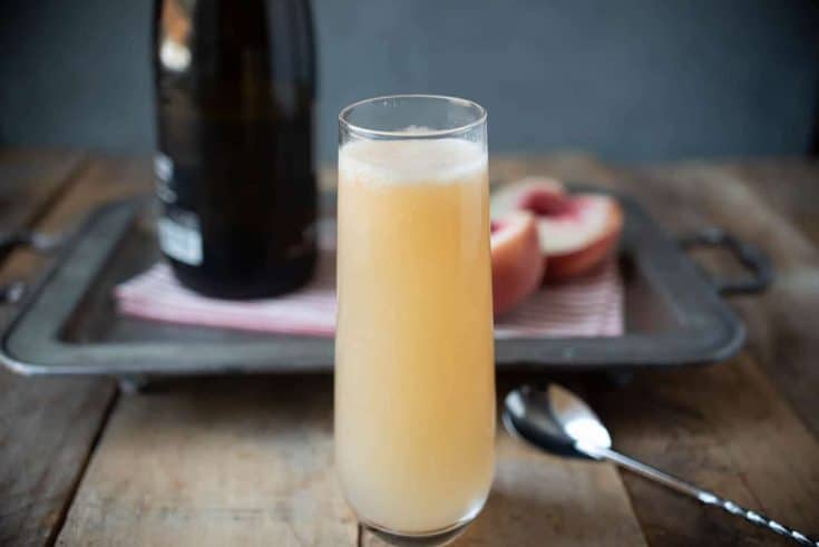 Prosecco and peach puree mixed in a stemless tall Champagne glass with a bottle of Prosecco and peach halves