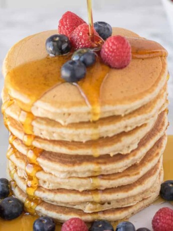 A large stack of pancakes topped with fruit pouring over syrup