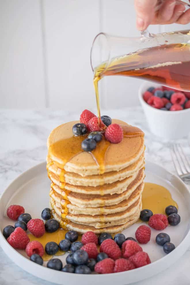 Pouring syrup of a large stack of pancakes surrounded with raspberries and blueberries