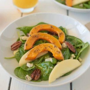 Sliced roasted butternut squash over spinach with sliced apples and pecans in a white bowl