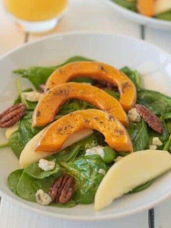 Sliced roasted butternut squash over spinach with sliced apples and pecans in a white bowl