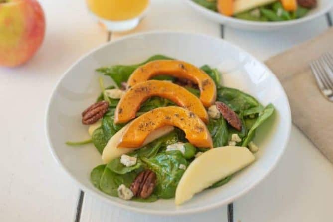 Sliced roasted butternut squash over spinach with sliced apples and pecans in a white bowl