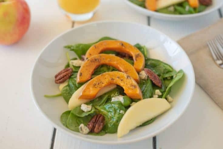 Sliced roasted butternut squash over spinach with sliced apples and pecans in a white bowl